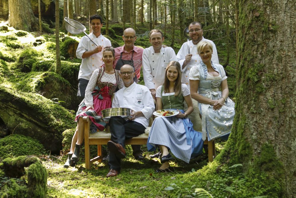 Männer und Frauen sitzen als Gruppe mit zubereiteten Gerichten im Wald.