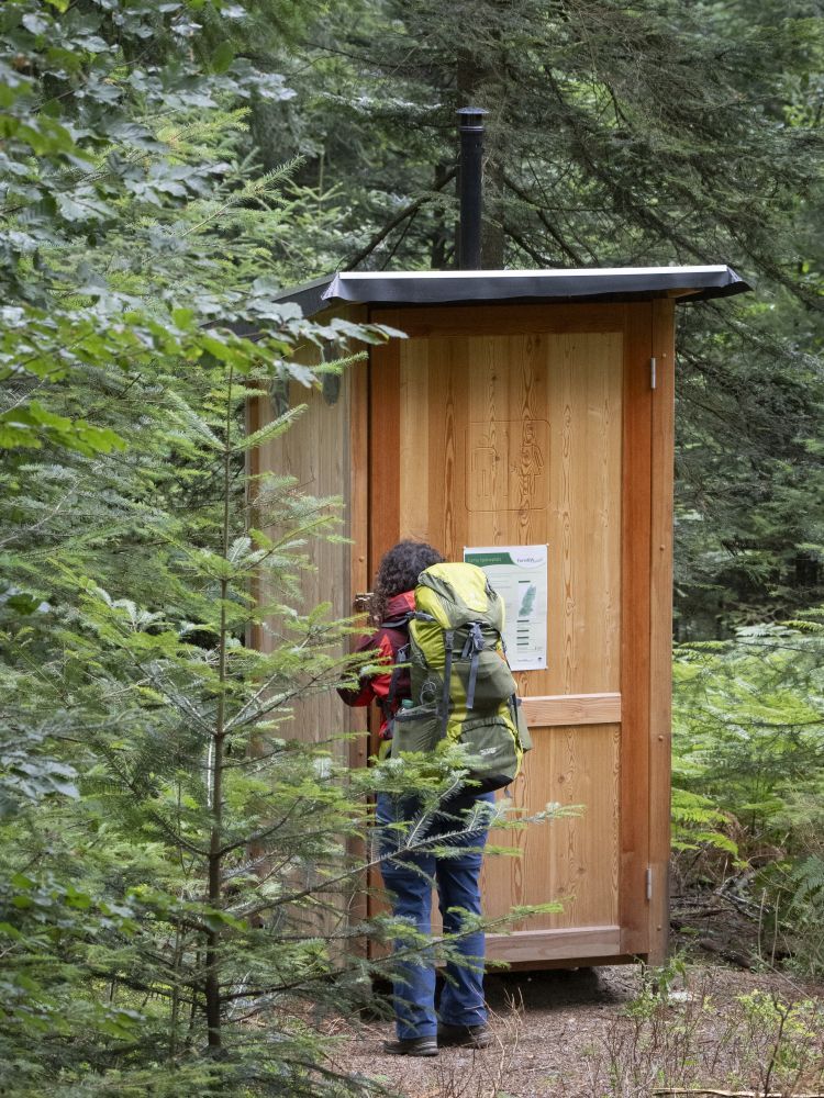 Eine Frau steht vor einem Toilettenhäuschen aus Holz, umgeben von Bäumen.