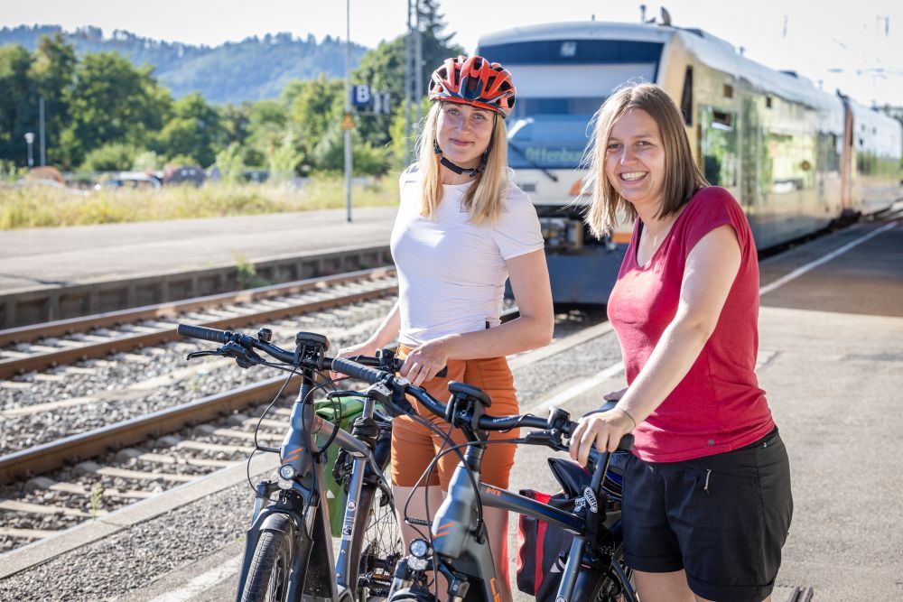 Zwei Frauen schieben ihr Fahhrad während im Hintergrund ein Zug am Bahnhof einfährt.