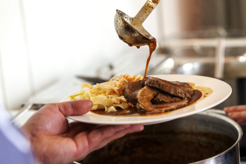 Bratensoße fließt von einem Schöpflöffel auf einen Teller mit Nudeln und Fleisch.