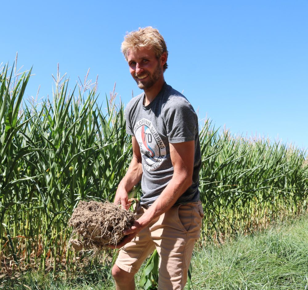 Ein Mann steht vor einem Maisfeld und hält eine Schaufel mit humusreicher Erde in der Hand.