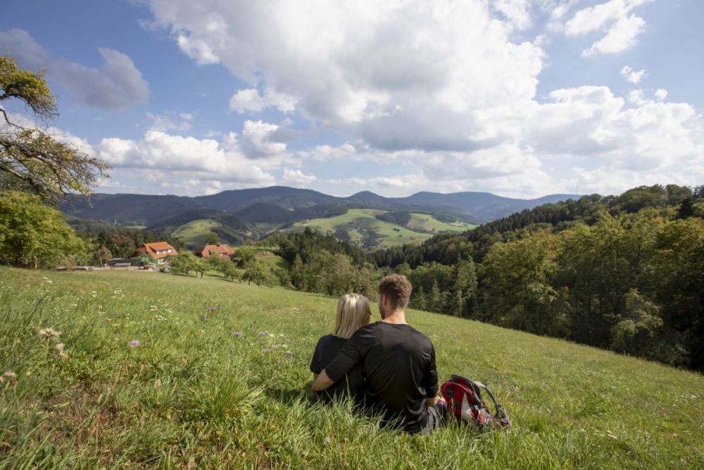 Zwei Menschen sitzen auf einer Wiese und schauen auf die Schwarzwälder Kulturlandschaft mit Weiden, Streuobstwiesen und Wald. Foto: Ulrike Klumpp